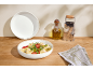 Nudelsalat in einer weißen Schüssel, mit einer Olivenölflasche und einer Reibe.