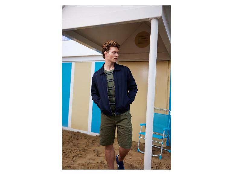 Mann in marineblauer Jacke, gestreiftem T-Shirt und Cargoshorts am Sandstrand.