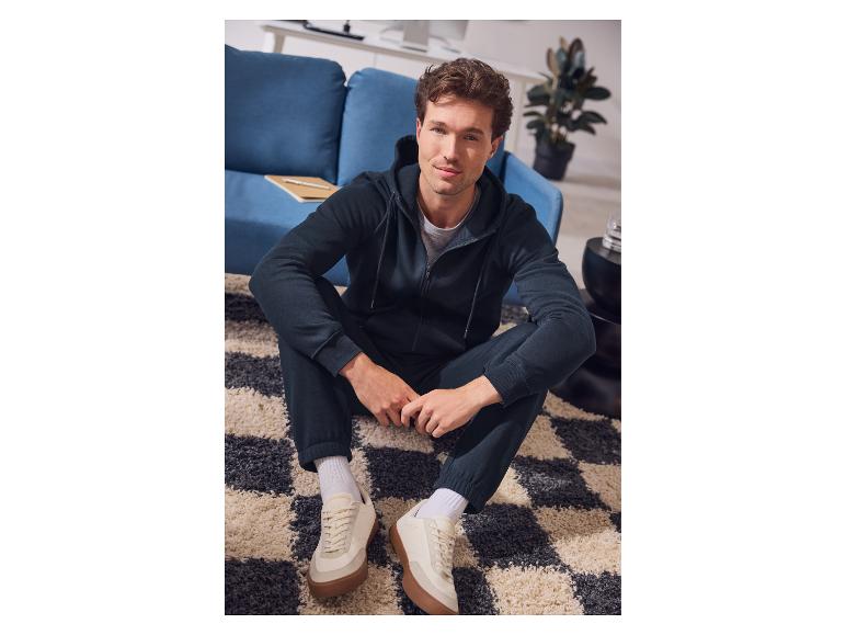 Mann in dunkelblauem Kapuzenpullover und Jogginghose sitzt auf einem Teppich.