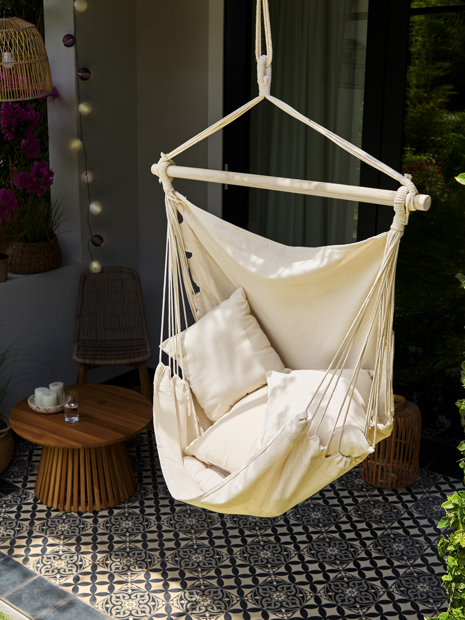 Cremefarbener Hängesessel mit Kissen, Holztisch und gemusterten Fliesen auf einem Balkon.
