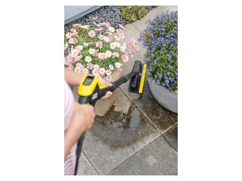 Frau reinigt Terrasse mit Hochdruckreiniger und Bürste, Blumen im Hintergrund.