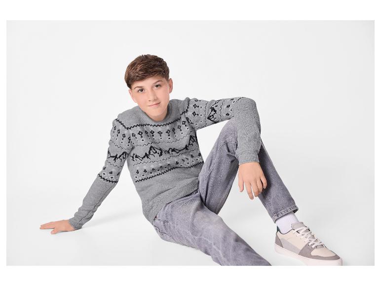 Junge in grauem gemustertem Strickpullover und grauen Jeans sitzt auf dem Boden.