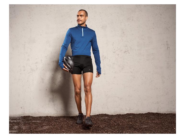 Ein Mann trägt ein blaues Oberteil und schwarze Radlerhose mit einem Helm in der Hand.