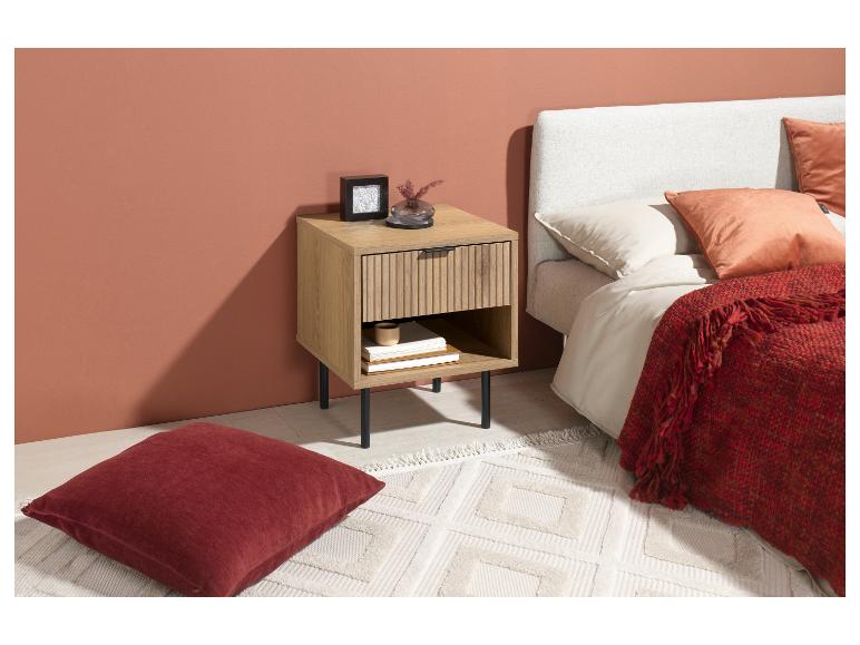 Nachttisch aus hellem Holz mit Schublade und Ablage, neben einem Bett mit roten Kissen und Decke.