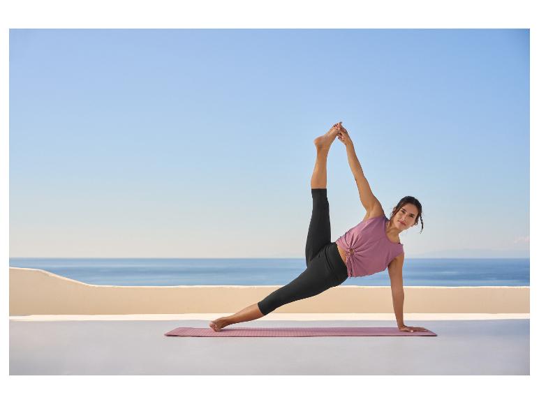 Frau in lila Top und schwarzen Leggings macht Yoga auf einer Matte auf einer Terrasse mit Meerblick.