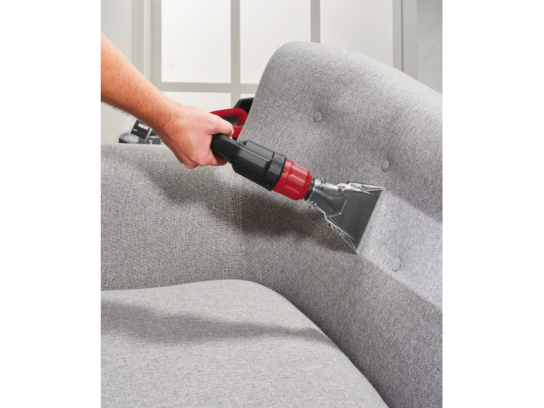 Mann reinigt ein graues Sofa mit einem rot-schwarzen Polsterreiniger.