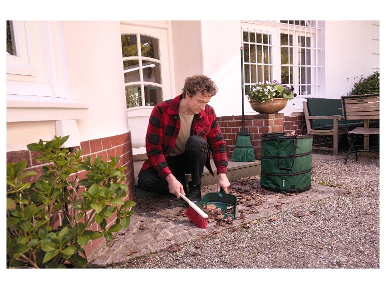 Mann kehrt Blätter mit Besen und Kehrschaufel in einen grünen Gartenabfallsack.
