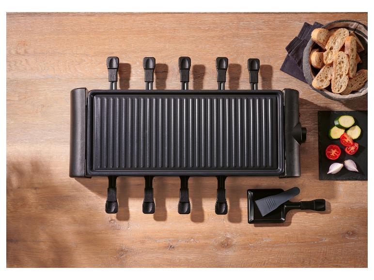 Raclette-Grill mit sechs Pfännchen, Brot und geschnittenem Gemüse auf einem Holztisch.