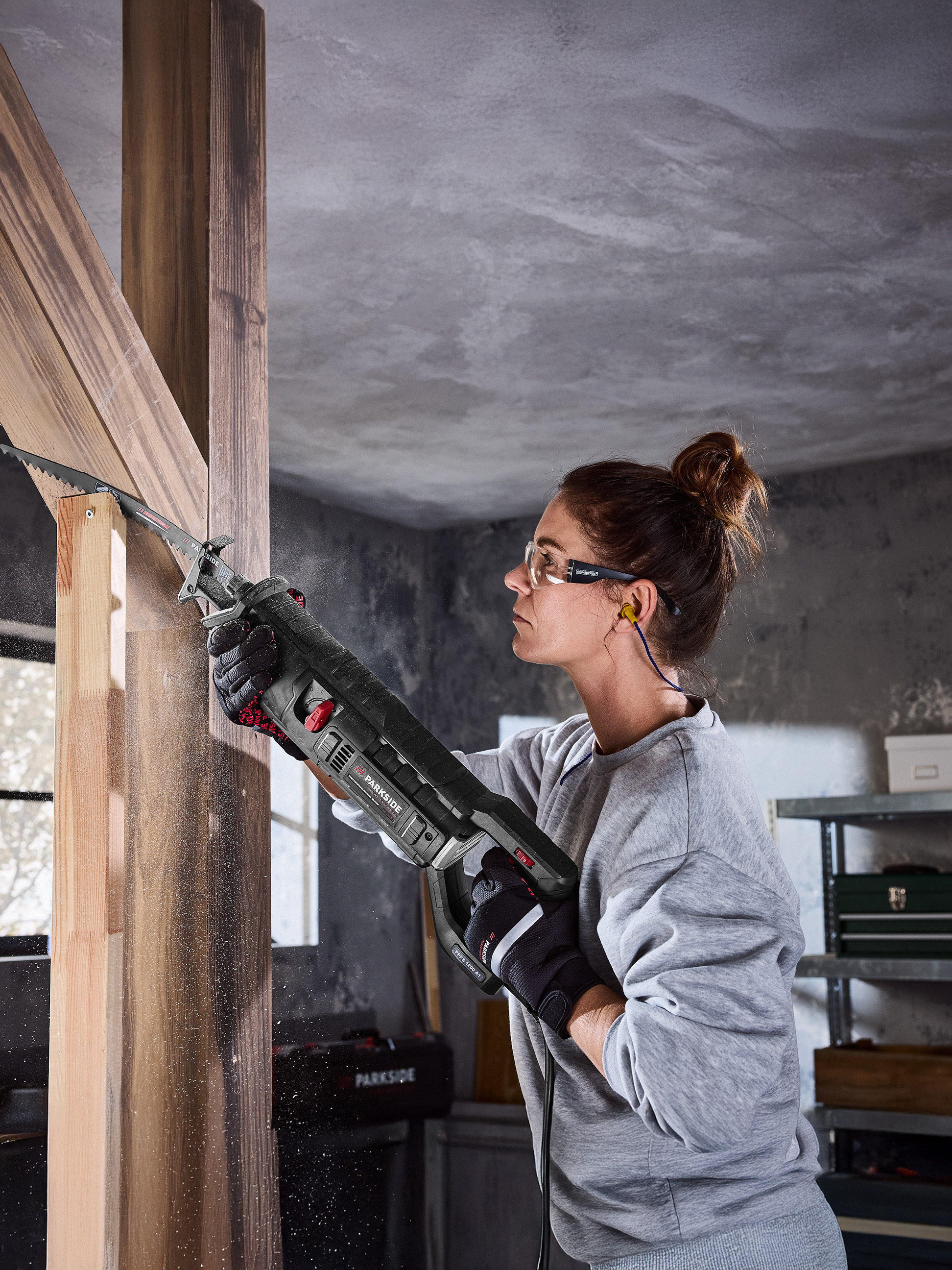 Frau benutzt Parkside Säbelsäge zum Sägen von Holzbalken, mit Schutzbrille und Gehörschutz.