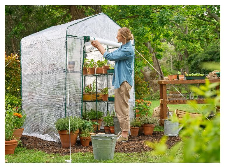 Frau öffnet ein kleines Gewächshaus mit Topfpflanzen und Gartengeräten.
