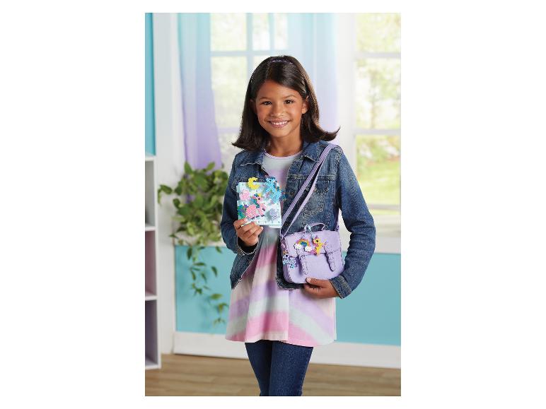 Lächelndes Mädchen hält ein SYLVANIAN FAMILIES Bastelset mit Einhorn-Perlen und eine lila Tasche mit Perlenanhängern.