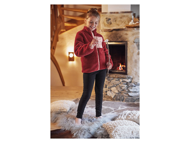 Ein Mädchen in einer roten Fleecejacke und schwarzen Leggings steht vor einem Kamin.