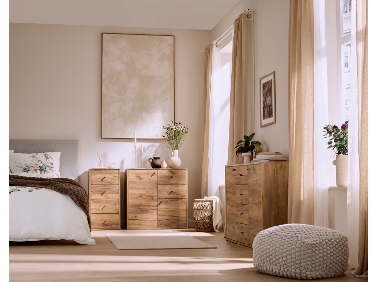 Schlafzimmer mit Holzkommoden, Bett mit Kopfteil und Zierkissen sowie beigen Vorhängen.