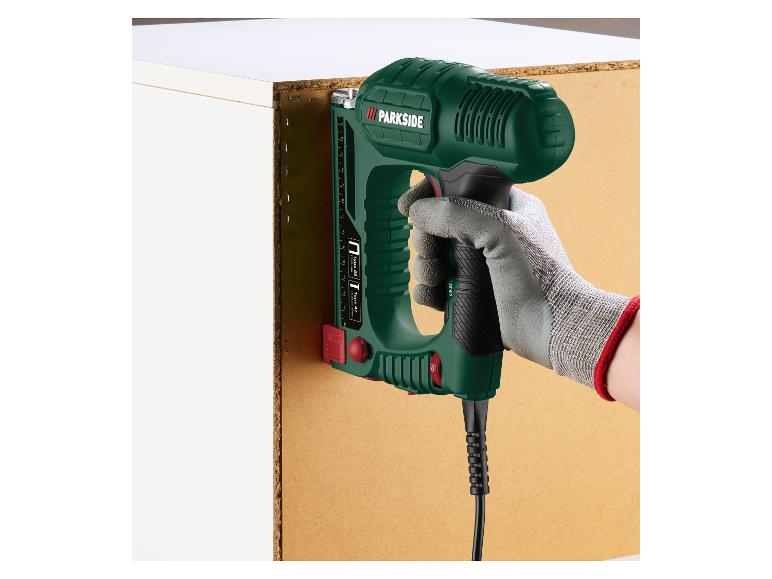 Hand hält einen Elektrotacker, der eine Holzplatte an einem weißen Schrank befestigt.