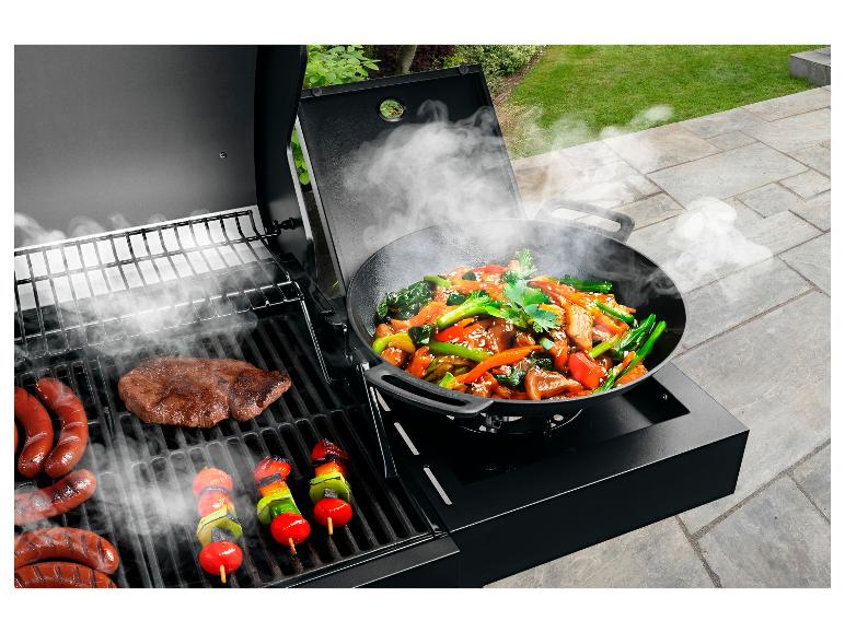 Grill mit Fleisch, Würstchen, Gemüsespießen und einer Wokpfanne mit asiatischem Gericht auf der Terrasse.