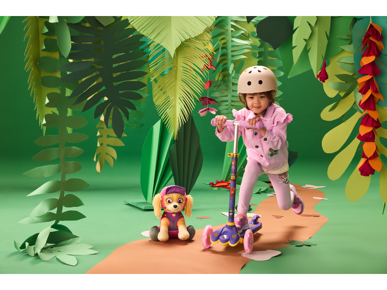 Mädchen mit Helm auf einem Roller, neben einem Plüschhund, in einer Dschungelkulisse.
