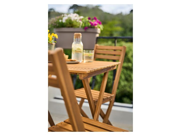 Auf einem Holzbalkontisch stehen eine Glaswasserflasche und Snacks, im Hintergrund Blumen und Grün.