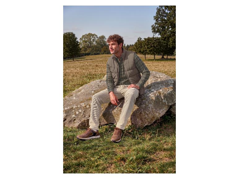 Mann in grüner Steppweste, kariertem Hemd und beiger Cordhose sitzt auf einem Felsen auf einem Feld.