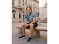 Mann in blauem Poloshirt und marineblauen Shorts sitzt auf einer Steinbank auf einem Stadtplatz.
