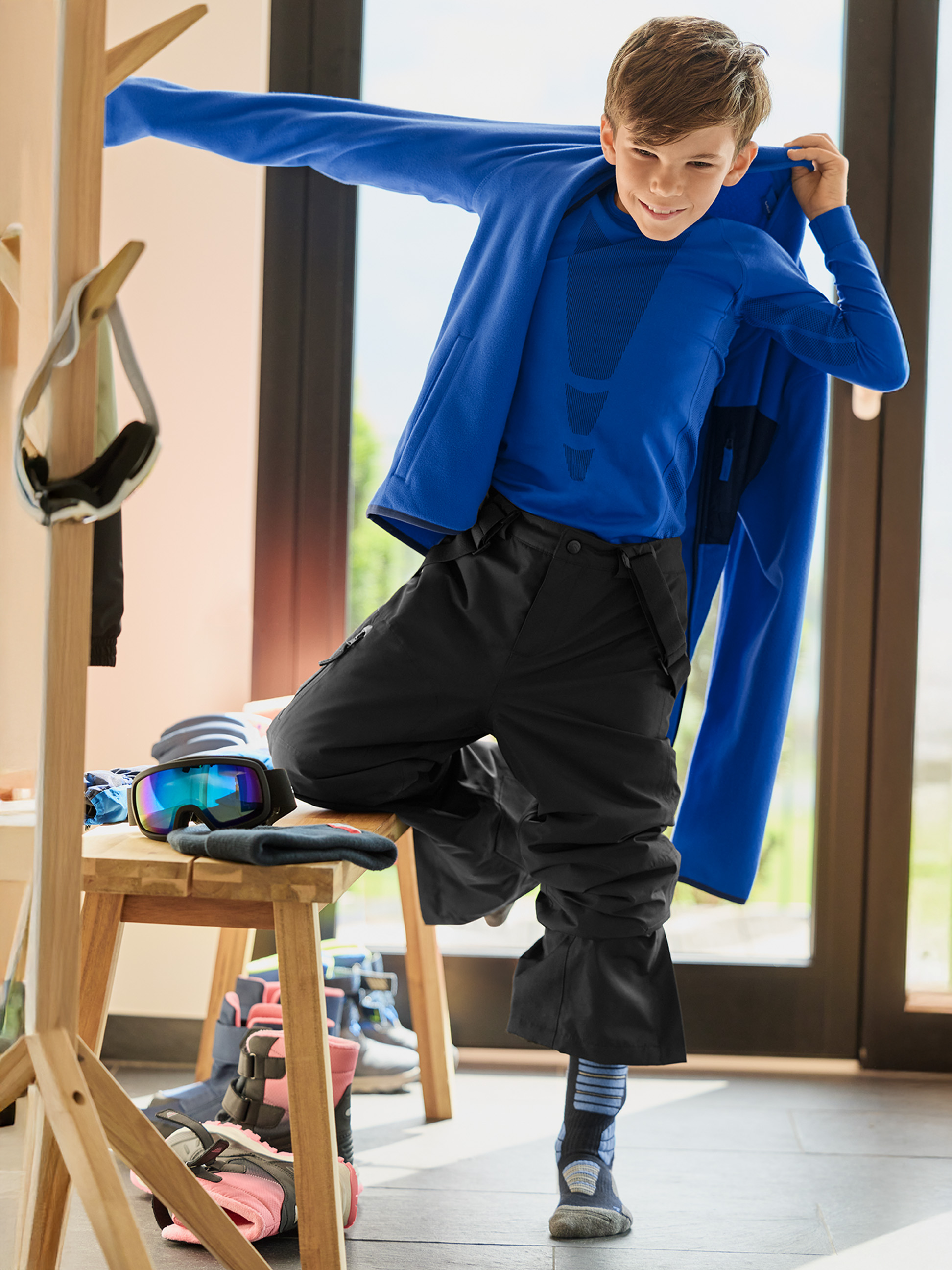 Ein lächelnder Junge in schwarzer Skihose und blauem Oberteil zieht eine blaue Jacke an.