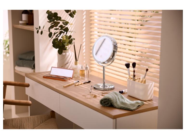 Schminktisch mit LED-Spiegel, Make-up-Pinseln und Kosmetik, mit Pflanze und Jalousien im Hintergrund.