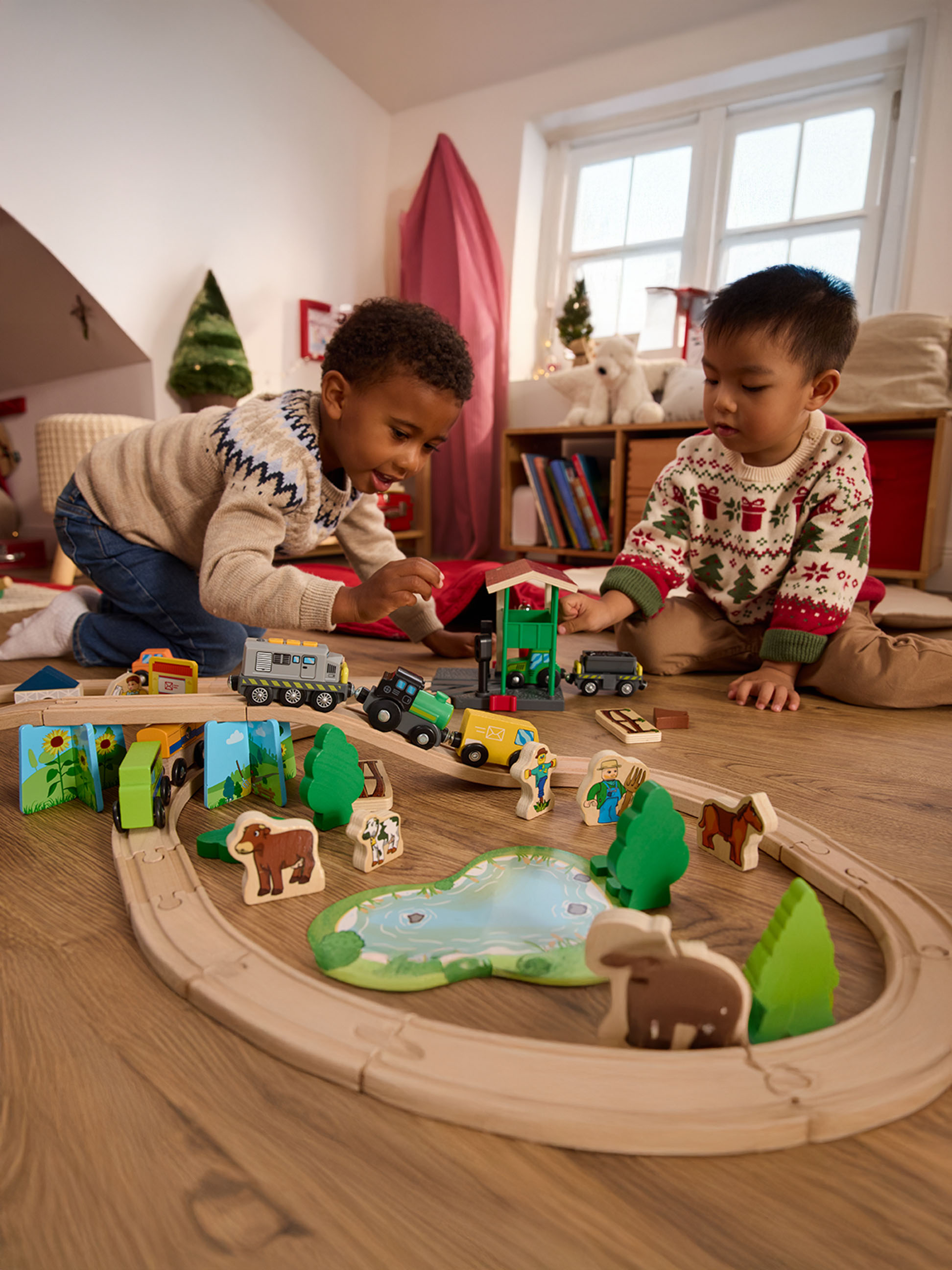 Zwei Jungen spielen mit einer Holzeisenbahn und Bauernhoftierfiguren auf einem Holzboden.