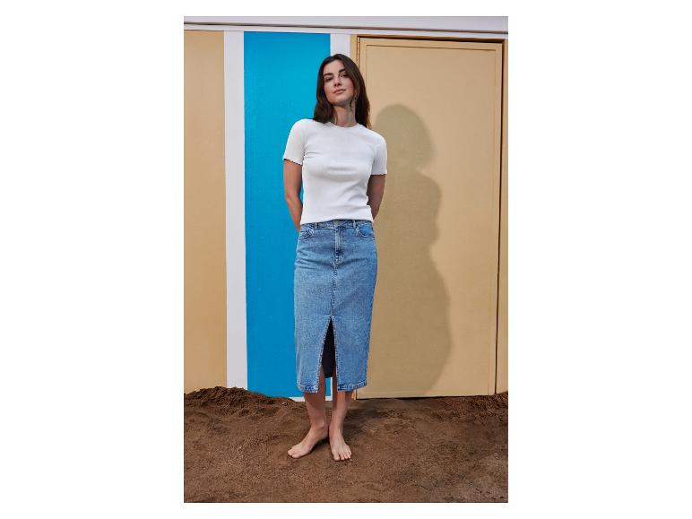 Frau in einem weißen gerippten T-Shirt und einem Jeans-Midirock, barfuß im Sand stehend.