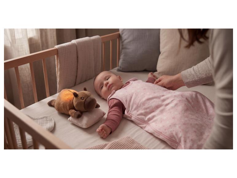 Baby in rosa Schlafsack im Kinderbett, mit leuchtendem Plüschtier daneben.