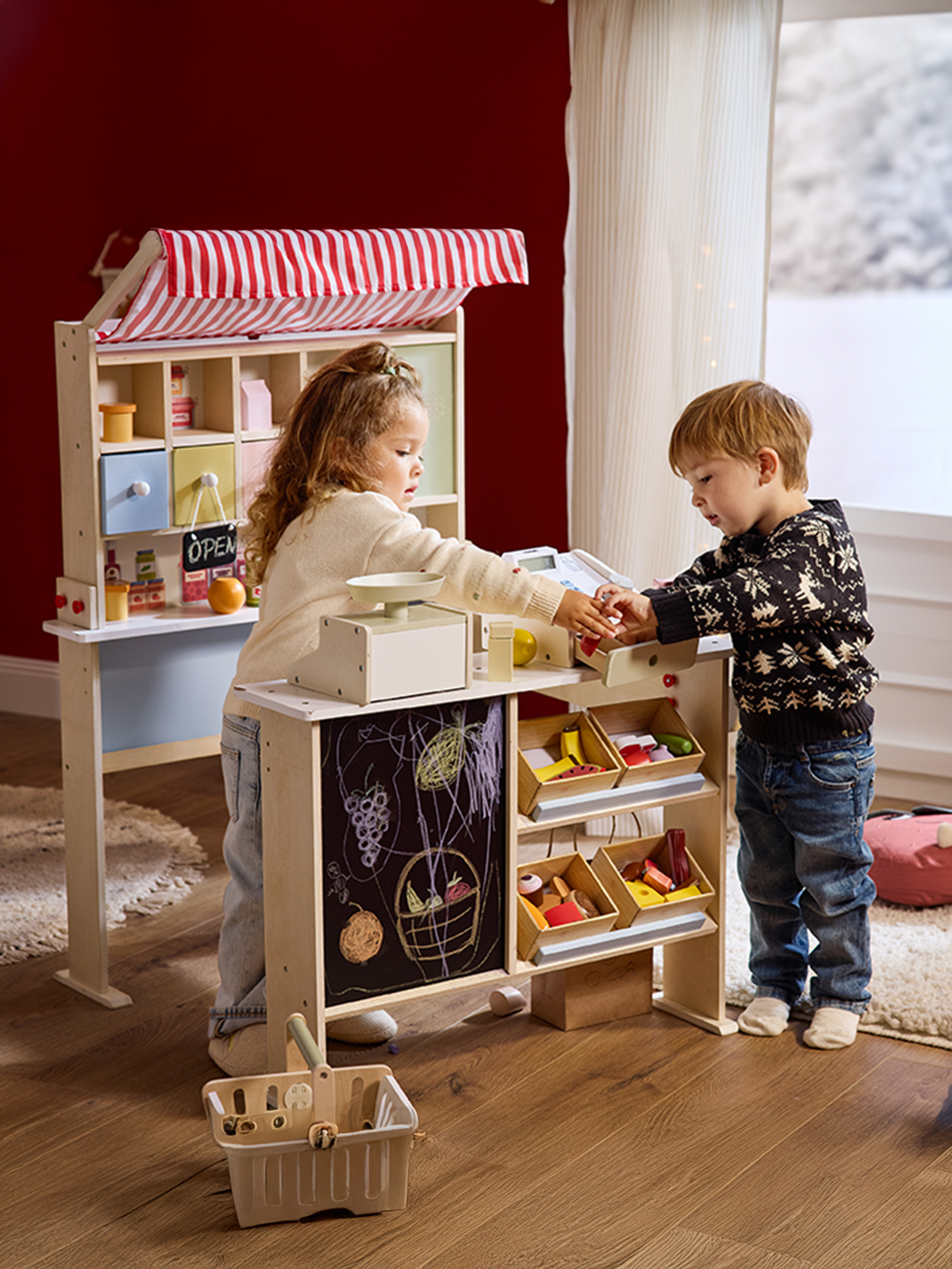 Zwei Kinder spielen mit einem Holzmarktstand, mit Kasse und Produkten.