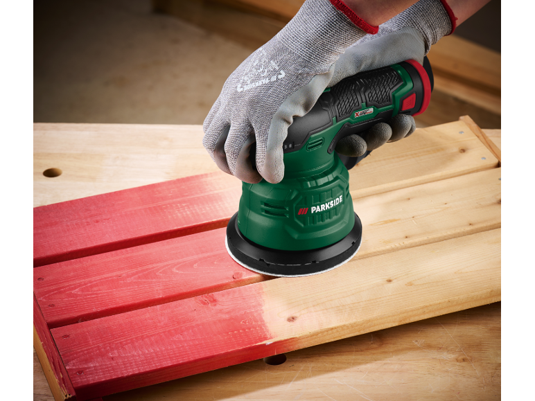 Eine behandschuhte Hand schleift ein rotes Holzbrett mit einem Exzenterschleifer.