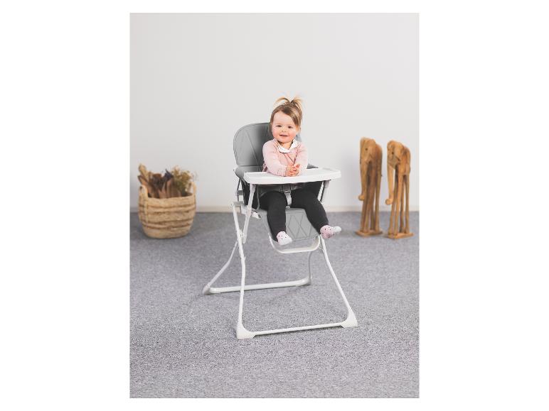 Ein lächelndes Baby sitzt in einem grau-weißen Hochstuhl auf einem grauen Teppich.