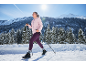 Frau in rosa Jacke und lila Leggings beim Nordic Walking in verschneiter Landschaft.