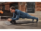 Ein Mann im blauen Trainingsanzug macht Unterarmstütz auf einer schwarzen Fitnessmatte mit einem Bauchroller.