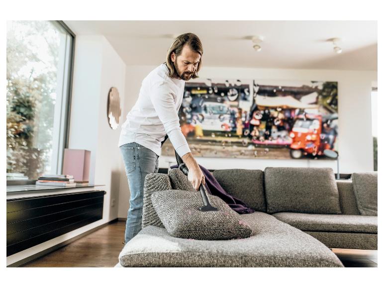 Mann reinigt ein graues Sofa mit einem Handstaubsauger.