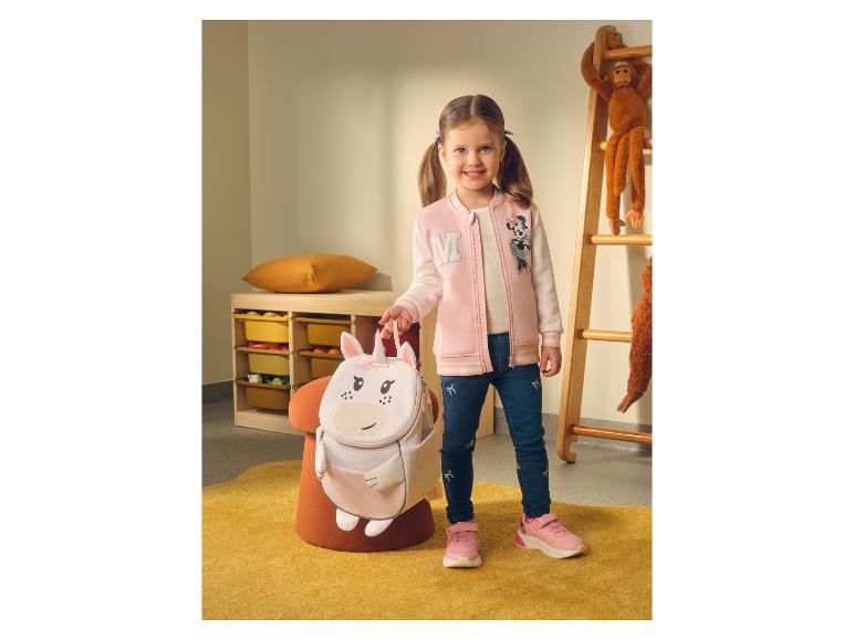 Lächelndes Mädchen in rosa Jacke und Jeans, hält einen Einhorn-Rucksack.