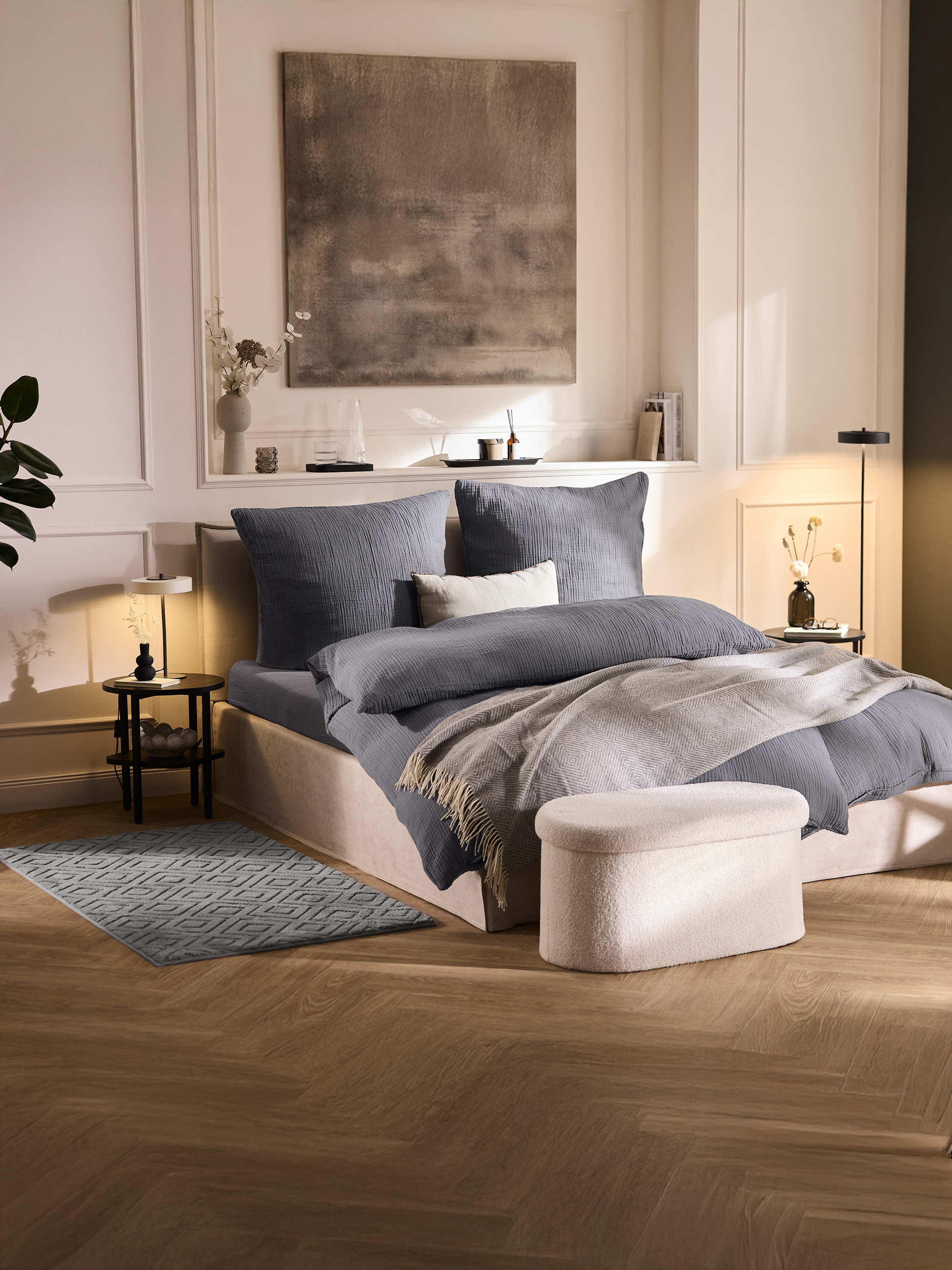 Schlafzimmer mit Bett mit grauer Bettwäsche, einem hellgrauen Überwurf und einem cremefarbenen Hocker.