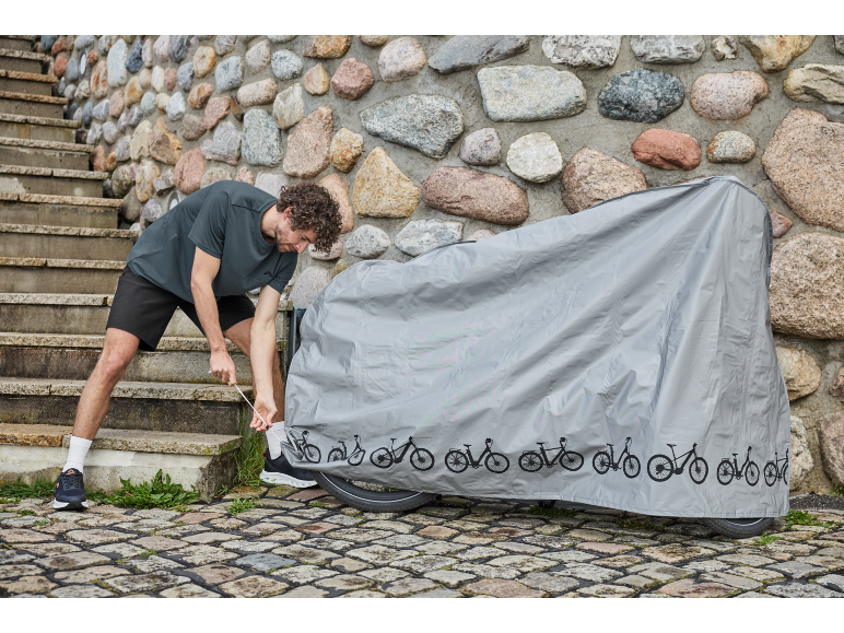 Ein Mann zieht eine graue Schutzhülle über ein Fahrrad neben einer Steinmauer.