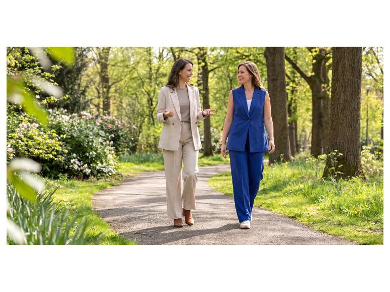 Zwei Frauen in eleganten Hosenanzügen gehen lächelnd durch einen Park.