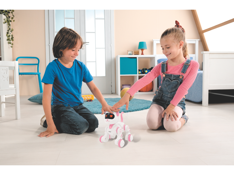 Zwei Kinder spielen mit einem weißen und rosa Roboterwelpen auf dem Boden.