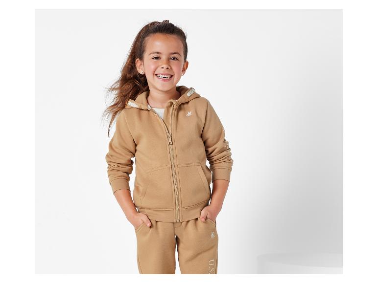 Ein Mädchen trägt einen beigen U.S. Polo Assn. Hoodie und Jogginghose.