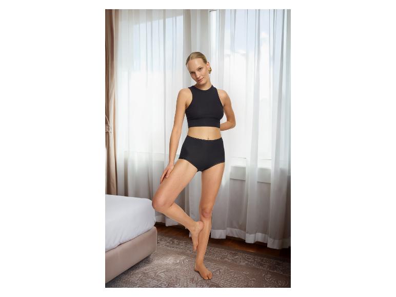 Frau in schwarzer Unterwäsche, Crop-Top und High-Waist-Slip, posiert in einem Schlafzimmer.