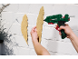 Hände halten eine Heißklebepistole und eine Holzfeder, die an eine weiße Wand geklebt wird.