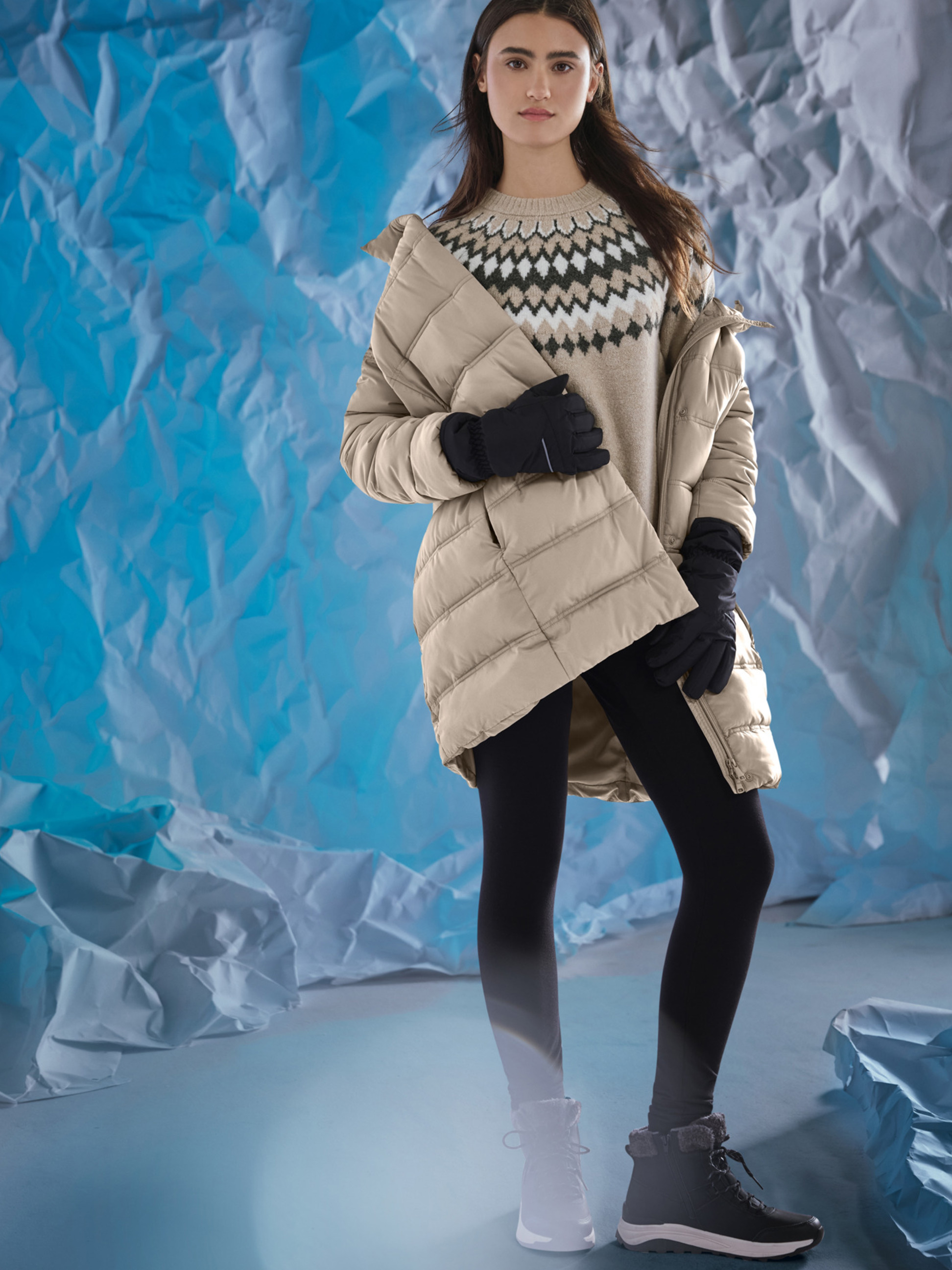 Frau in beiger Jacke, gemustertem Pullover, schwarzen Leggings und Winterstiefeln, vor blauem Hintergrund.