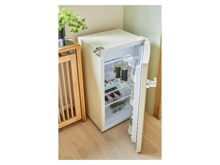 Offener cremefarbener Minikühlschrank mit Getränken und Essen, in einer Ecke neben einem Holztisch platziert.