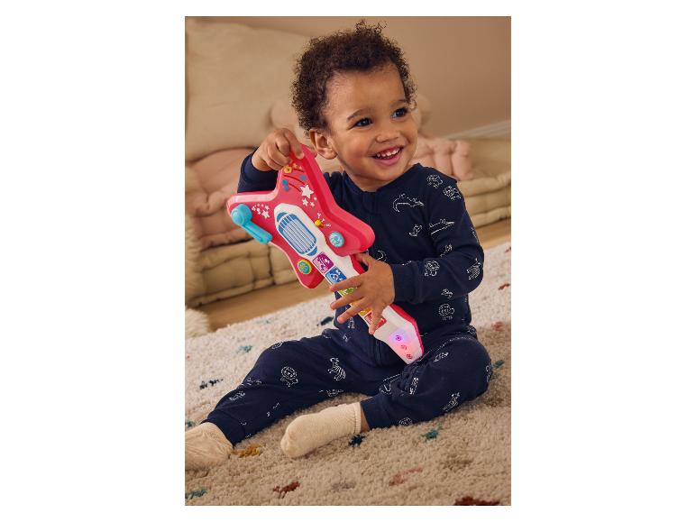 Ein lächelndes Baby in blauem Strampler spielt mit einer rosa Spielzeuggitarre.