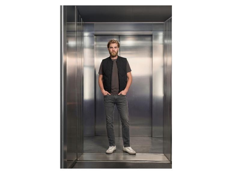 Mann in Weste im Aufzug, trägt Jeans und T-Shirt.