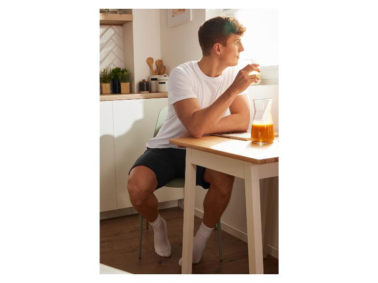 Mann in weißem T-Shirt und Shorts genießt Orangensaft zu Hause.