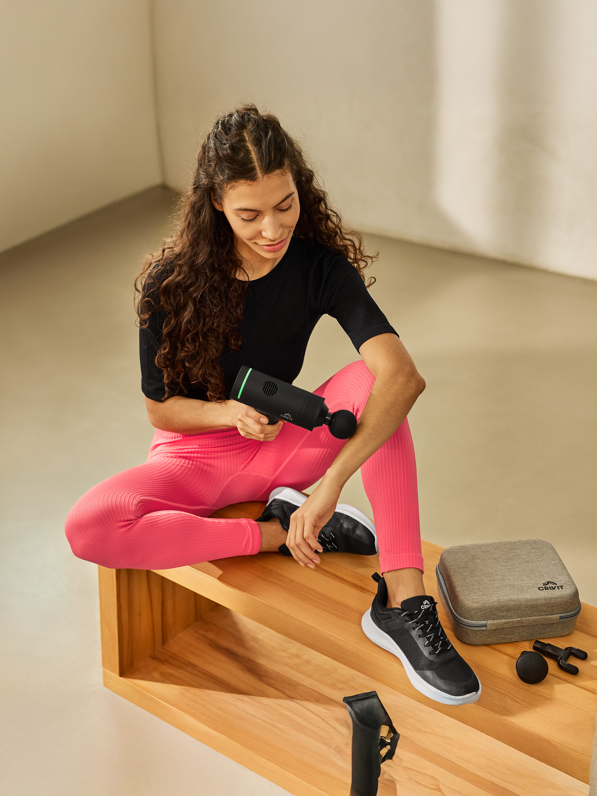 Frau benutzt Massagepistole, sitzt auf Holzbank, daneben Etui und Zubehör.