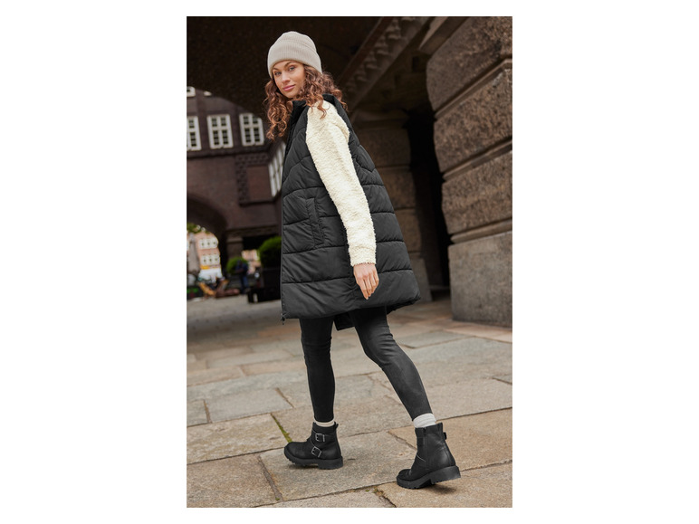 Eine Frau trägt eine schwarze gesteppte Weste, schwarze Hose und schwarze Stiefel.