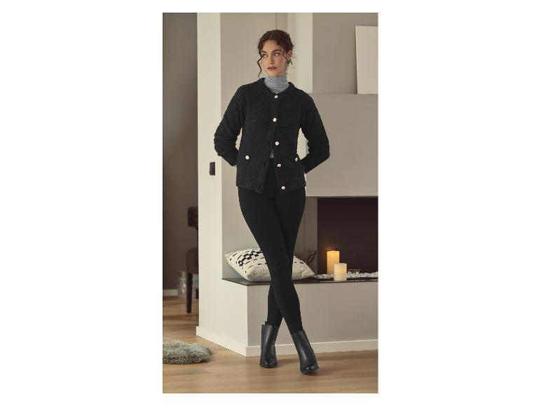 Frau in schwarzer Strickjacke, schwarzen Leggings und schwarzen Stiefeln, steht an einem Kamin.